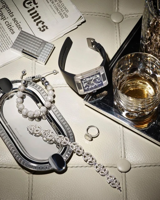 Wristwatch, jewelry, and barware are arranged on a tufted surface with a newspaper, lighter, and silver trays under studio lighting.