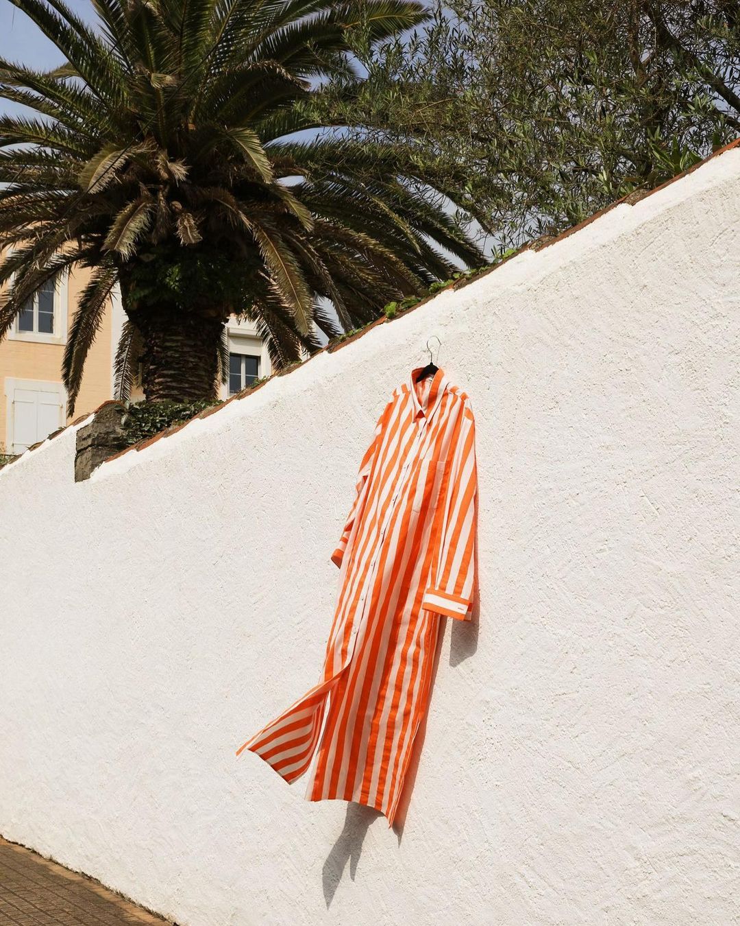 Orange and white striped dress hanging on a white textured wall with a palm tree and other greenery in the background on a sunny day.