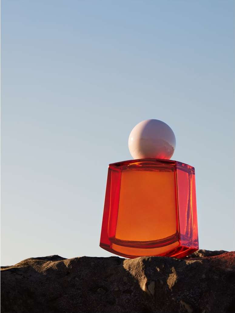 Still life shot of an orange glass perfume bottle with a white spherical cap, sitting on a textured rock surface against a light blue sky.