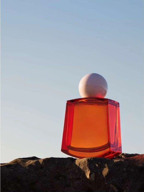 Still life shot of an orange glass perfume bottle with a white spherical cap, sitting on a textured rock surface against a light blue sky.