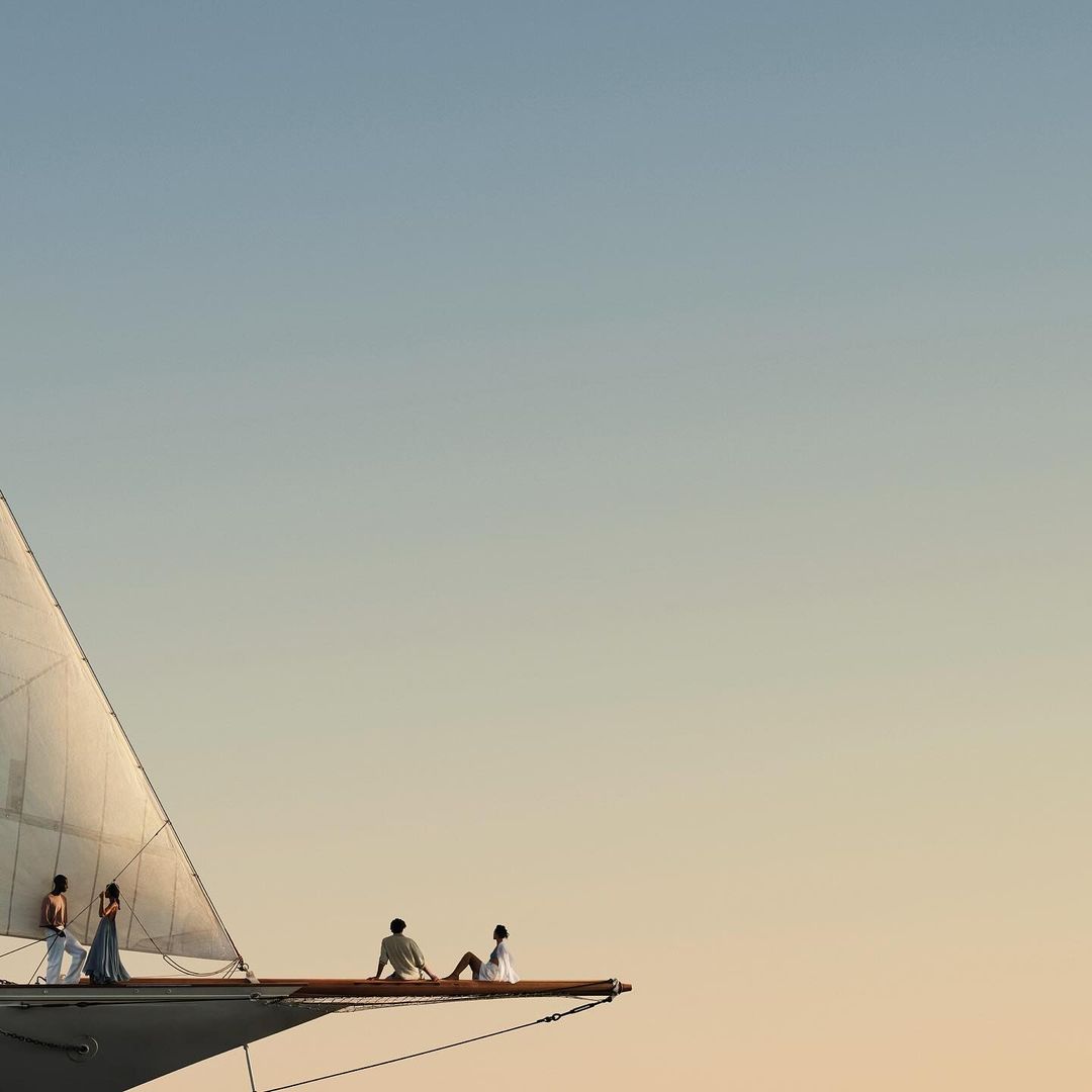 Four people sit on the bow of a large sailboat against a clear, gradient sky at dusk. The sail is visible on the left of the image.