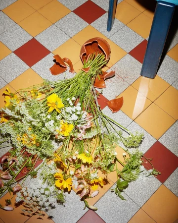 Still life featuring a bouquet of wildflowers scattered on checkered tiles beside a broken clay vase, with a blue chair leg visible.