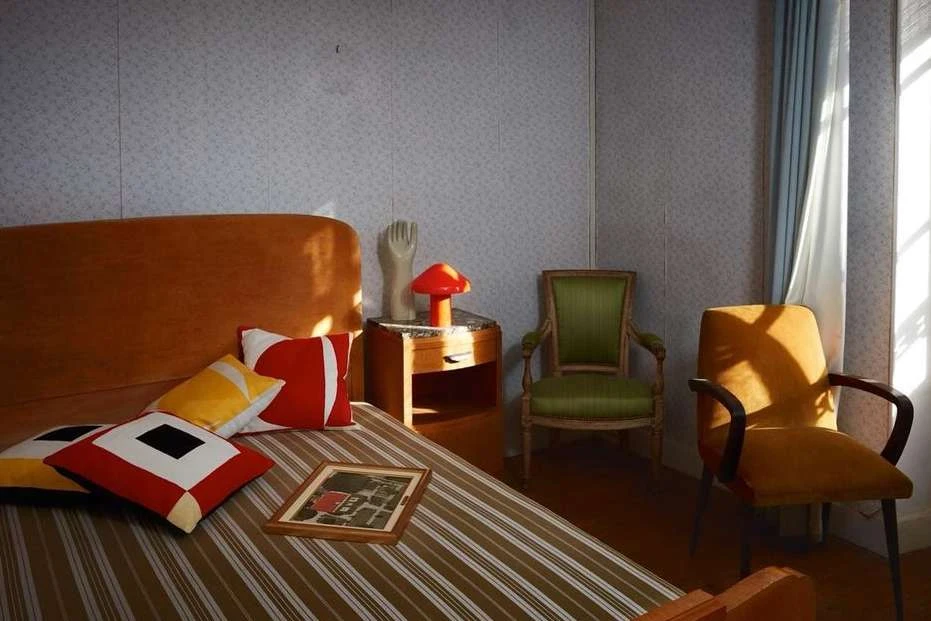Bedroom interior featuring a bed with geometric pillows, vintage chairs, a nightstand with a lamp, and a retro wallpaper design.