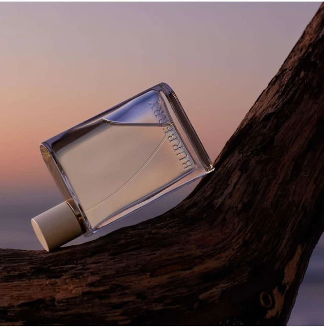 Burberry perfume bottle on a rustic wood surface, set against a soft gradient sky with orange and pink hues in an outdoor setting.