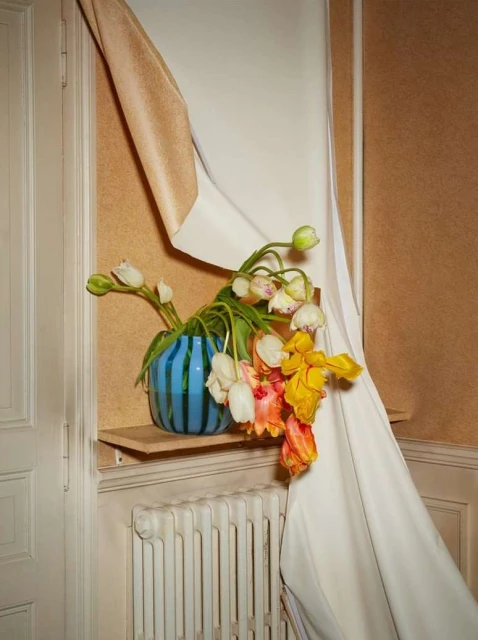 A blue striped vase holds a bouquet of white, peach, and yellow tulips indoors, against a beige wall and white curtain backdrop.