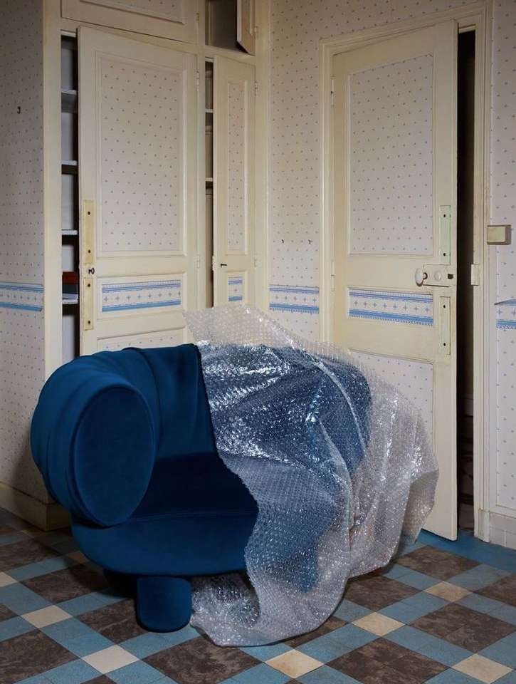Blue modern armchair partially covered in bubble wrap in a room with blue-and-white patterned wallpaper and doors.
