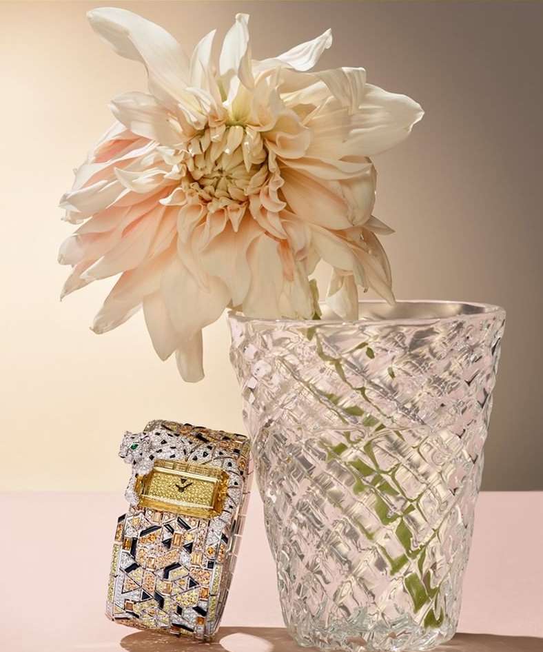 Still life of a luxury diamond watch next to a decorative crystal vase with a pastel flower against a graduated neutral background.