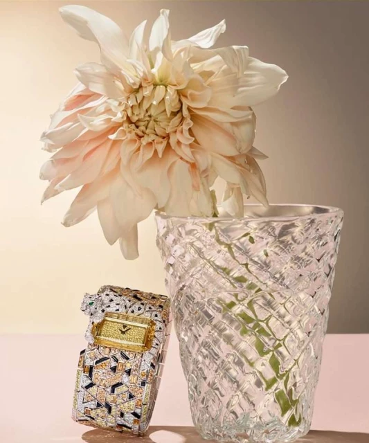 Still life of a luxury diamond watch next to a decorative crystal vase with a pastel flower against a graduated neutral background.