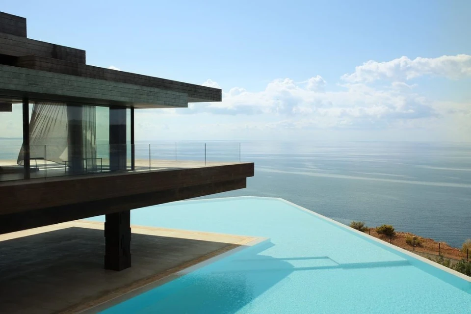 Modern luxury architecture showcases a minimalist villa with an infinity pool overlooking the ocean under a bright sky with light clouds.