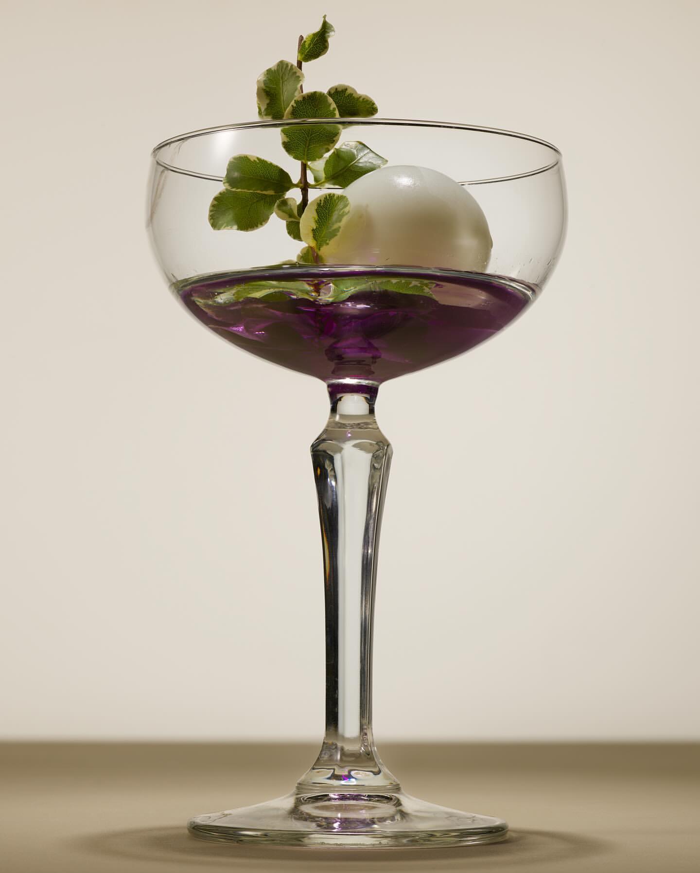 Elegant coupe glass filled with purple liquid, topped with a white sphere and a sprig of green leaves, set against a cream background.