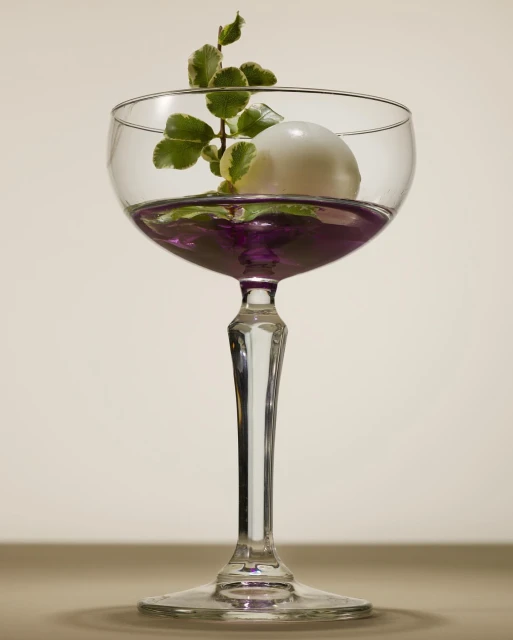 Elegant coupe glass filled with purple liquid, topped with a white sphere and a sprig of green leaves, set against a cream background.