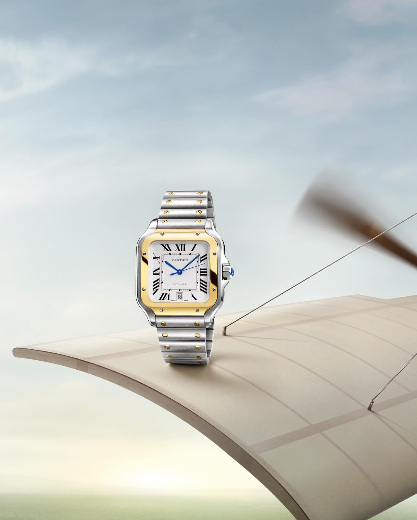 Luxury wristwatch with gold and silver band on the wing of an antique airplane against a blue sky backdrop, studio product shot.