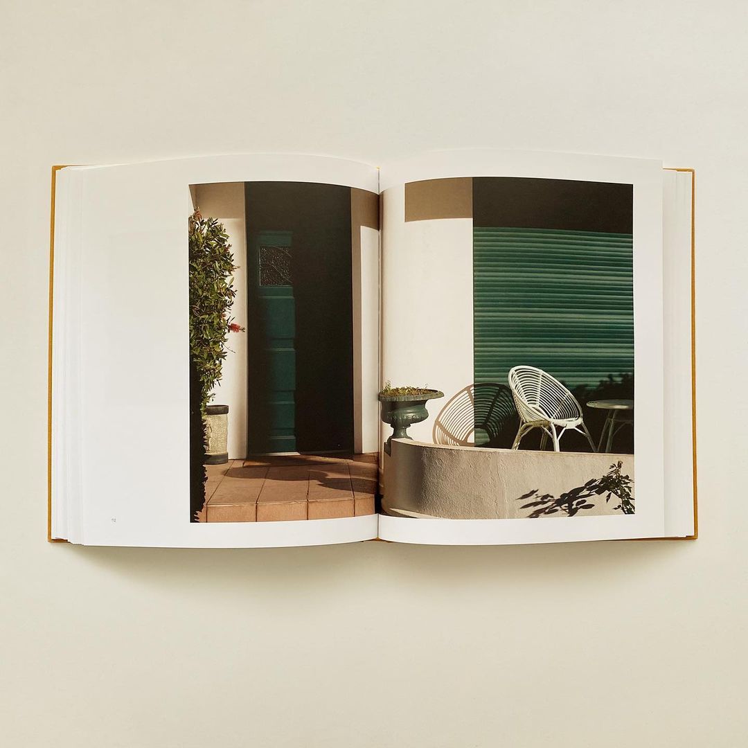 Open book shows a photo of an exterior home entrance with a dark door, planters, a low concrete wall, and chairs on a tile surface.