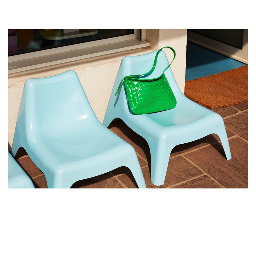 Bright green purse on light blue plastic chair on a brown tile floor. Two chairs are visible, outside on a sunny day.