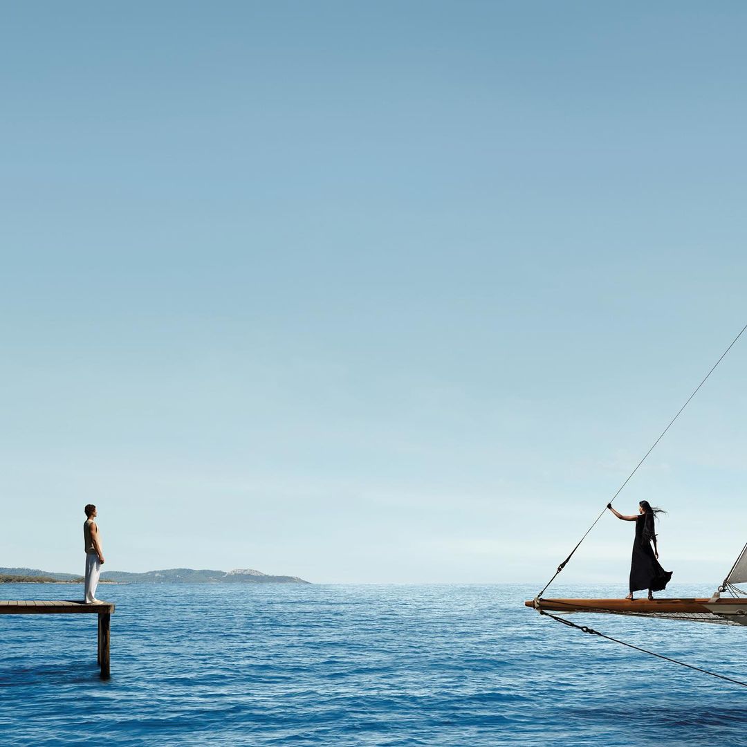 A person standing on a wooden pier looks across the open blue sea toward a woman standing on the bow of a sailboat.