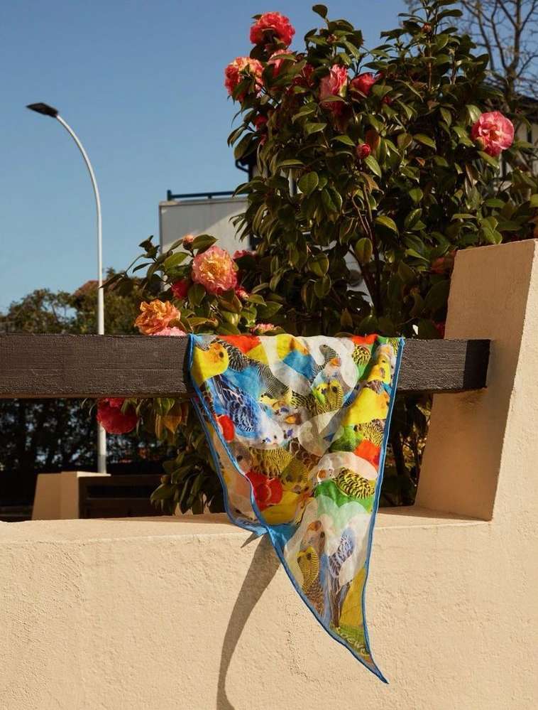 Colorful scarf with bird pattern draped on a wooden rail, set against a building, flowers, and a blue sky.
