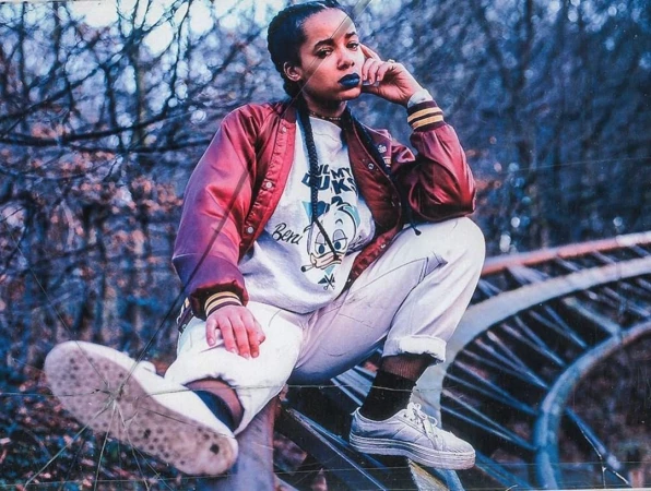 A young adult woman in a red bomber jacket sits on a metal structure outdoors, with a forest and blue sky in the background.