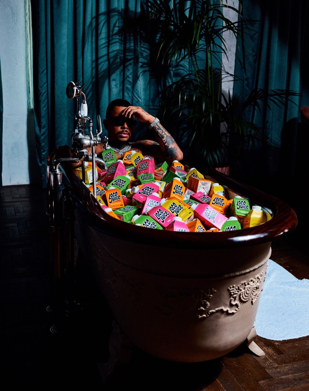 Adult male sitting in a brown clawfoot bathtub filled with colorful juice boxes, with a teal curtain and green plant in the background.