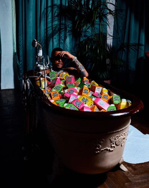 Adult male sitting in a brown clawfoot bathtub filled with colorful juice boxes, with a teal curtain and green plant in the background.