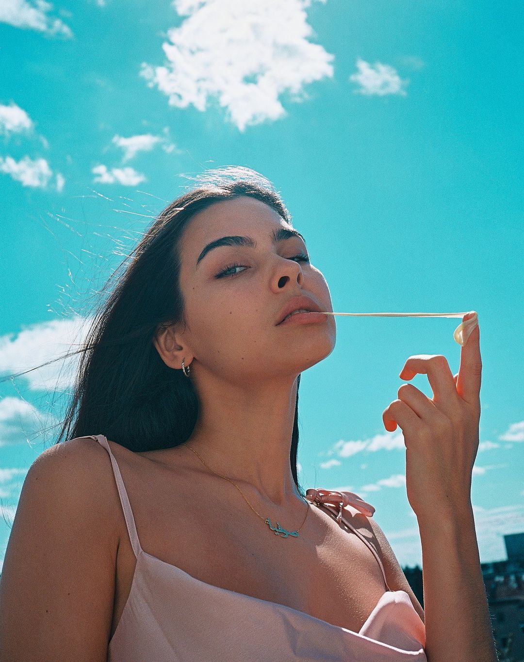 A young woman poses outdoors against a bright blue sky while stretching a piece of gum between her mouth and hand.