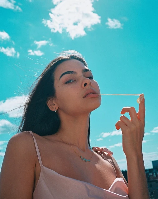 A young woman poses outdoors against a bright blue sky while stretching a piece of gum between her mouth and hand.