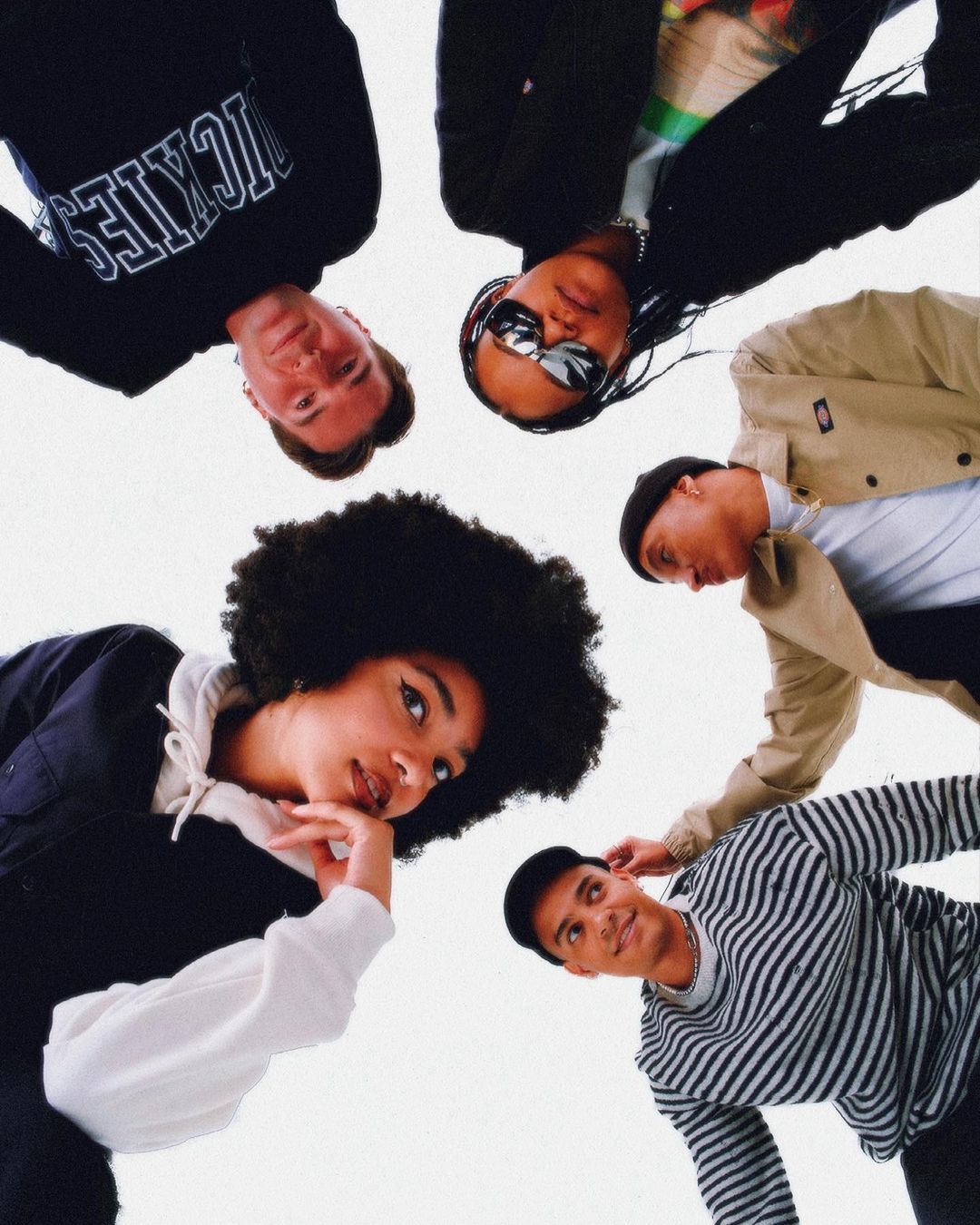 Eye-level group shot of five diverse young adults in casual clothing. Friends pose together looking down, showcasing casual style and unity.
