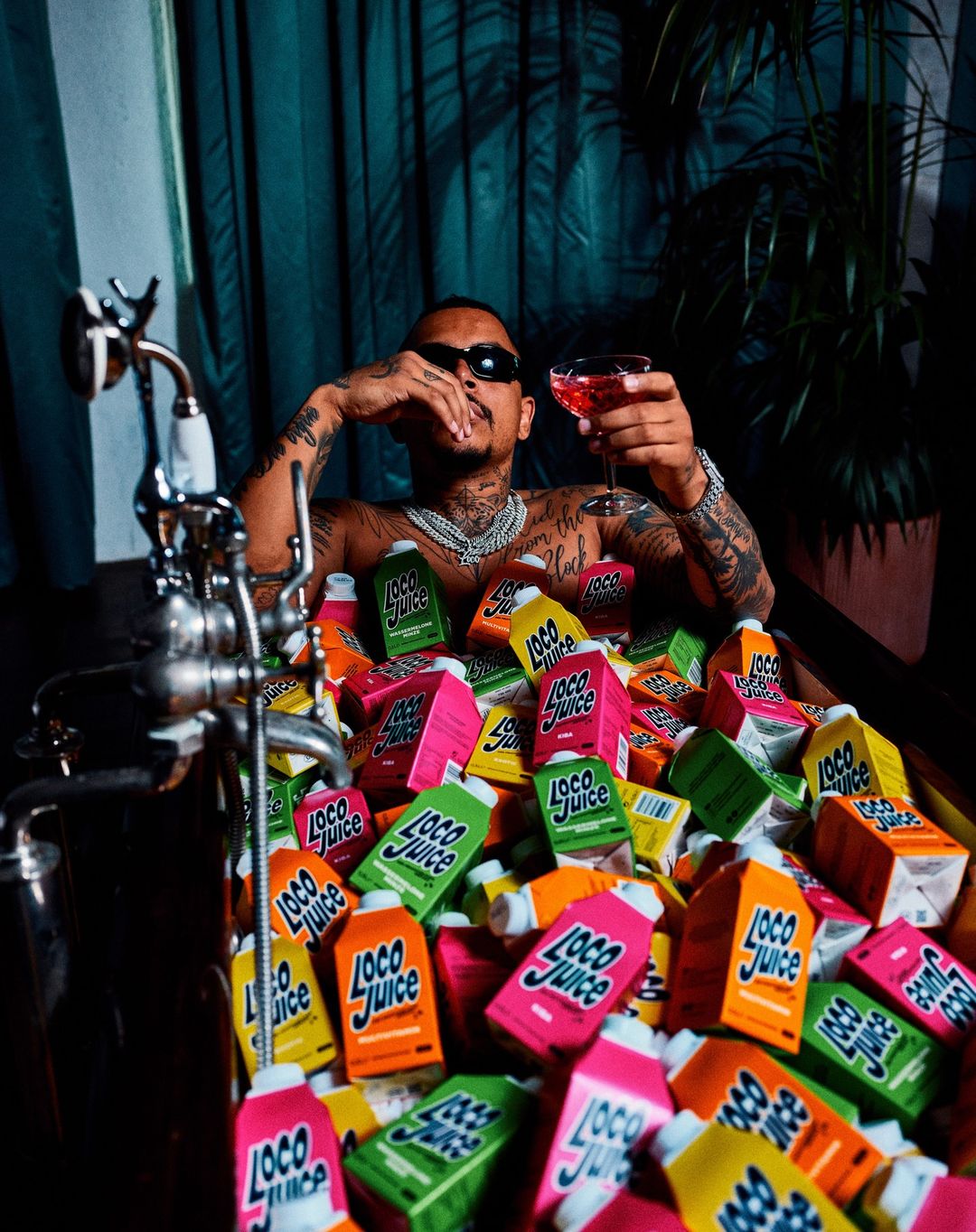Adult male with sunglasses sitting in a tub filled with Loco Juice cartons, holding a cocktail, against a curtain background.