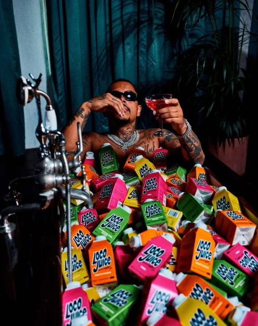 Adult male with sunglasses sitting in a tub filled with Loco Juice cartons, holding a cocktail, against a curtain background.