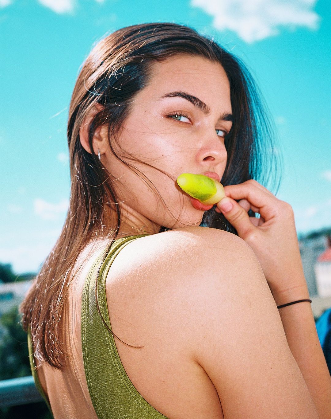 Portrait of a young woman in a green top enjoying a yellow and green popsicle against a blue sky with soft white clouds.