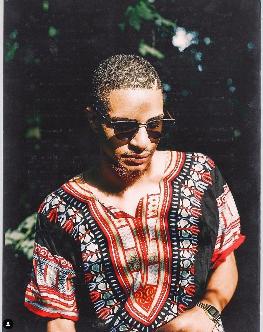 An adult male with short hair and sunglasses poses outdoors wearing an African-print shirt and a watch on his left wrist.