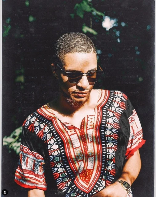 An adult male with short hair and sunglasses poses outdoors wearing an African-print shirt and a watch on his left wrist. 