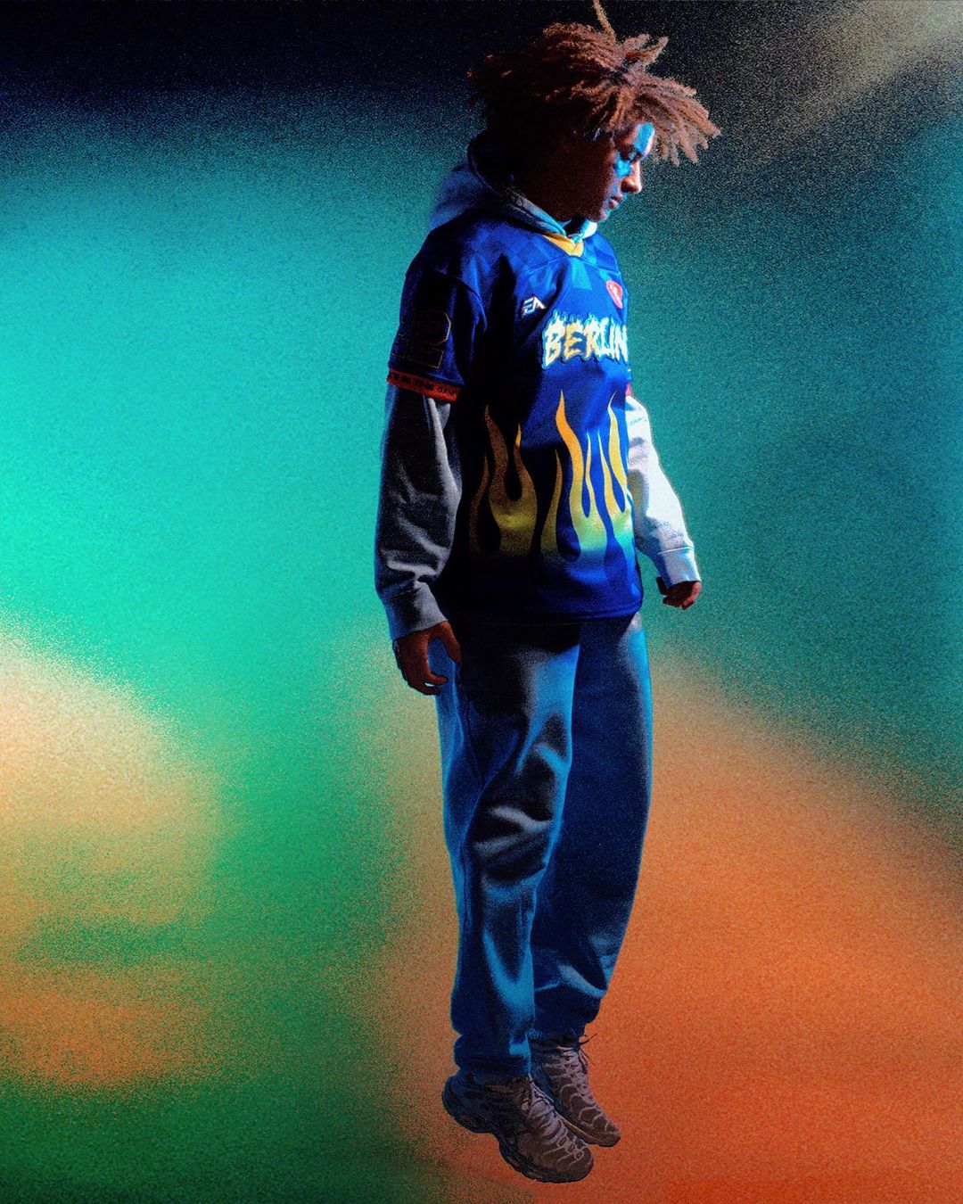 Full-body shot of a teenager wearing a blue sports jersey with flame graphics and grey sweats, set against a colorful blended backdrop.