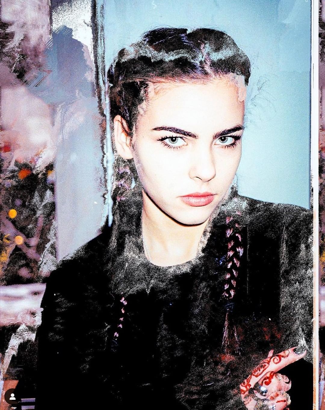 A young woman with braided hair poses indoors wearing a dark fur coat. Her green eyes are striking and she has designs on her hand.