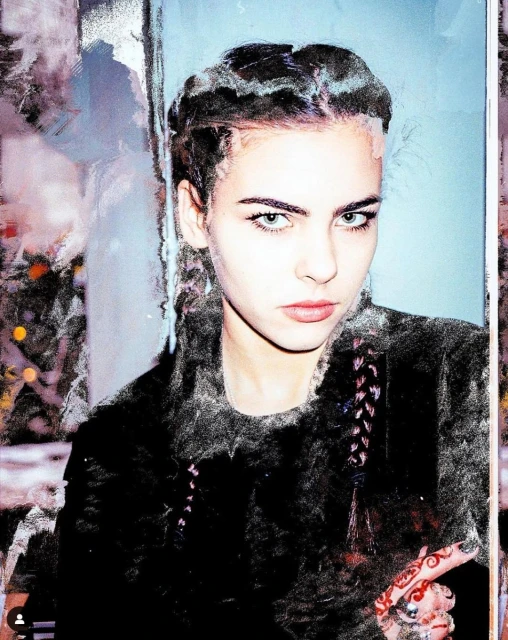 A young woman with braided hair poses indoors wearing a dark fur coat. Her green eyes are striking and she has designs on her hand.