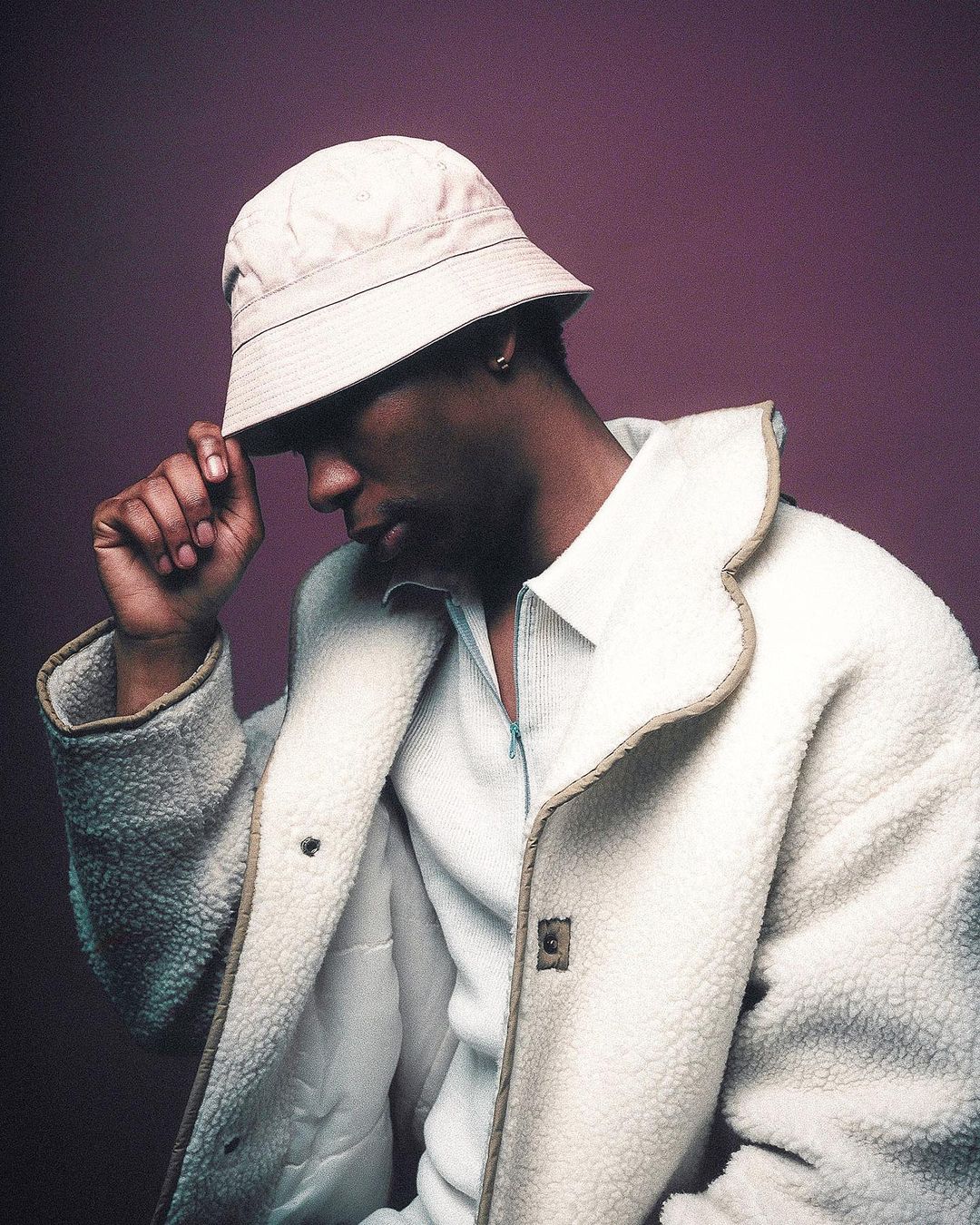 Portrait of a stylish black man in a cream-colored bucket hat and sherpa coat against a purple background in a studio setting.
