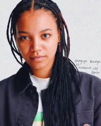 Portrait of a young woman with braided black hair wearing a dark canvas jacket over a white t-shirt, photographed by Roberto Brundo.