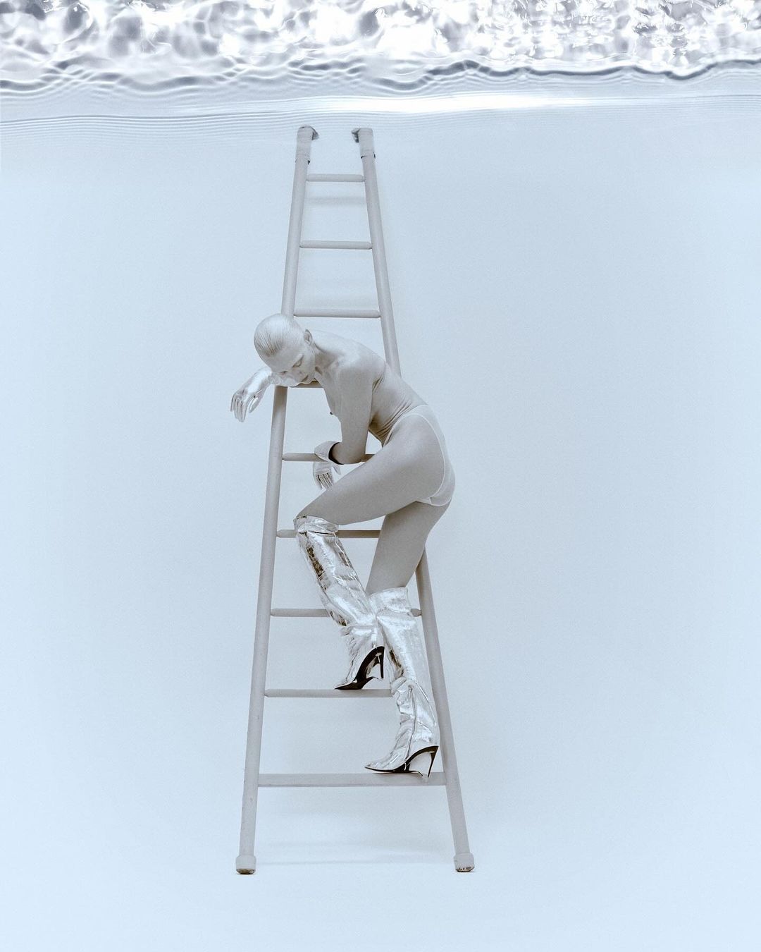 A woman in silver paint and matching boots climbs a silver ladder against a light blue backdrop with water ripples overhead.