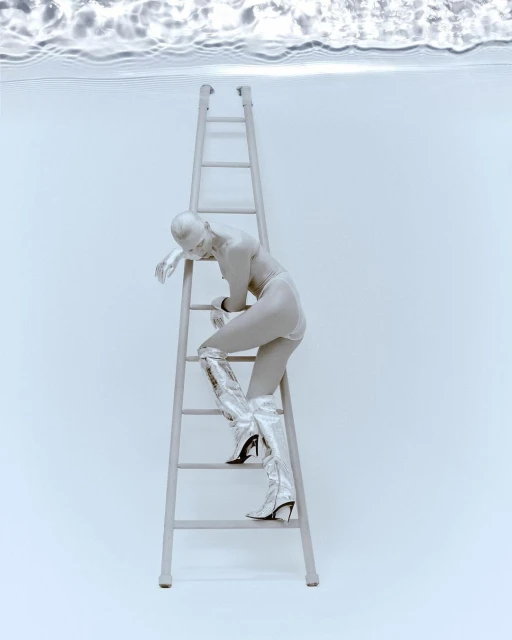 A woman in silver paint and matching boots climbs a silver ladder against a light blue backdrop with water ripples overhead.