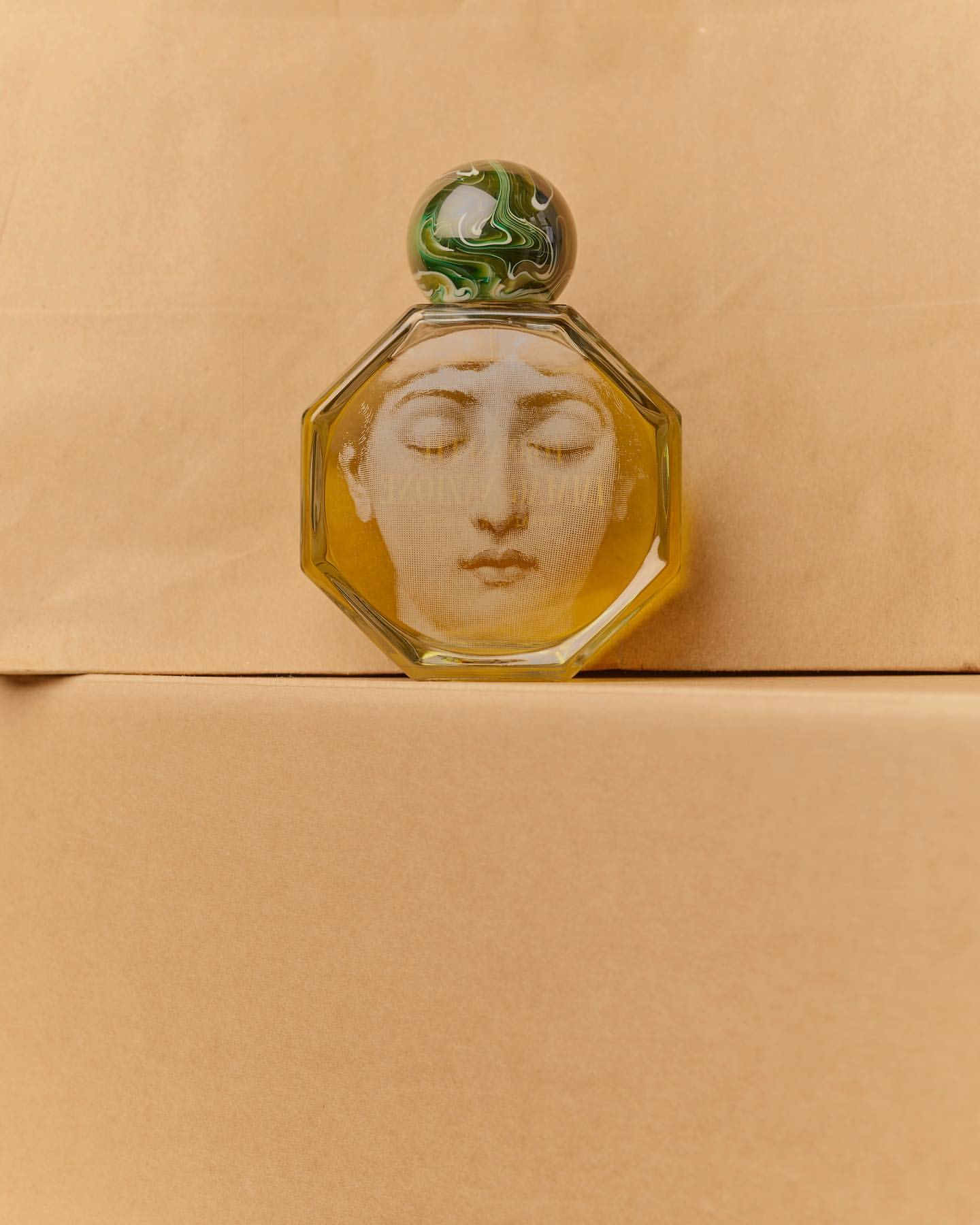 Close-up of an octagonal perfume bottle with a face design and a marbled green and white stopper, set against a beige backdrop.