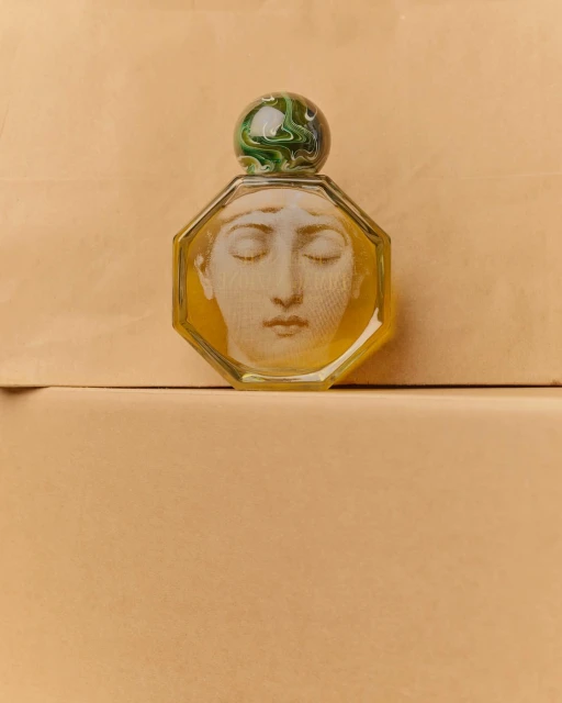 Close-up of an octagonal perfume bottle with a face design and a marbled green and white stopper, set against a beige backdrop.