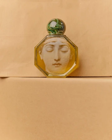 Close-up of an octagonal perfume bottle with a face design and a marbled green and white stopper, set against a beige backdrop.