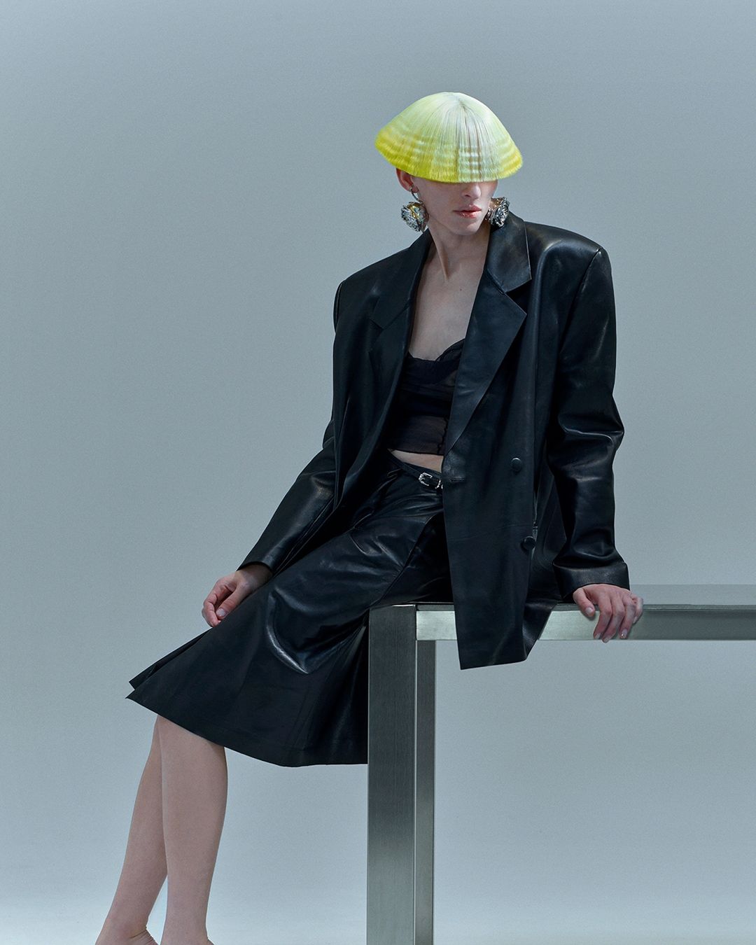 A person with short hair wearing a black leather jacket, skirt, and a sculptural hat sitting on a table against a grey background.