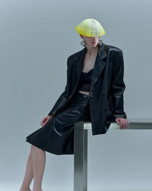 A person with short hair wearing a black leather jacket, skirt, and a sculptural hat sitting on a table against a grey background.