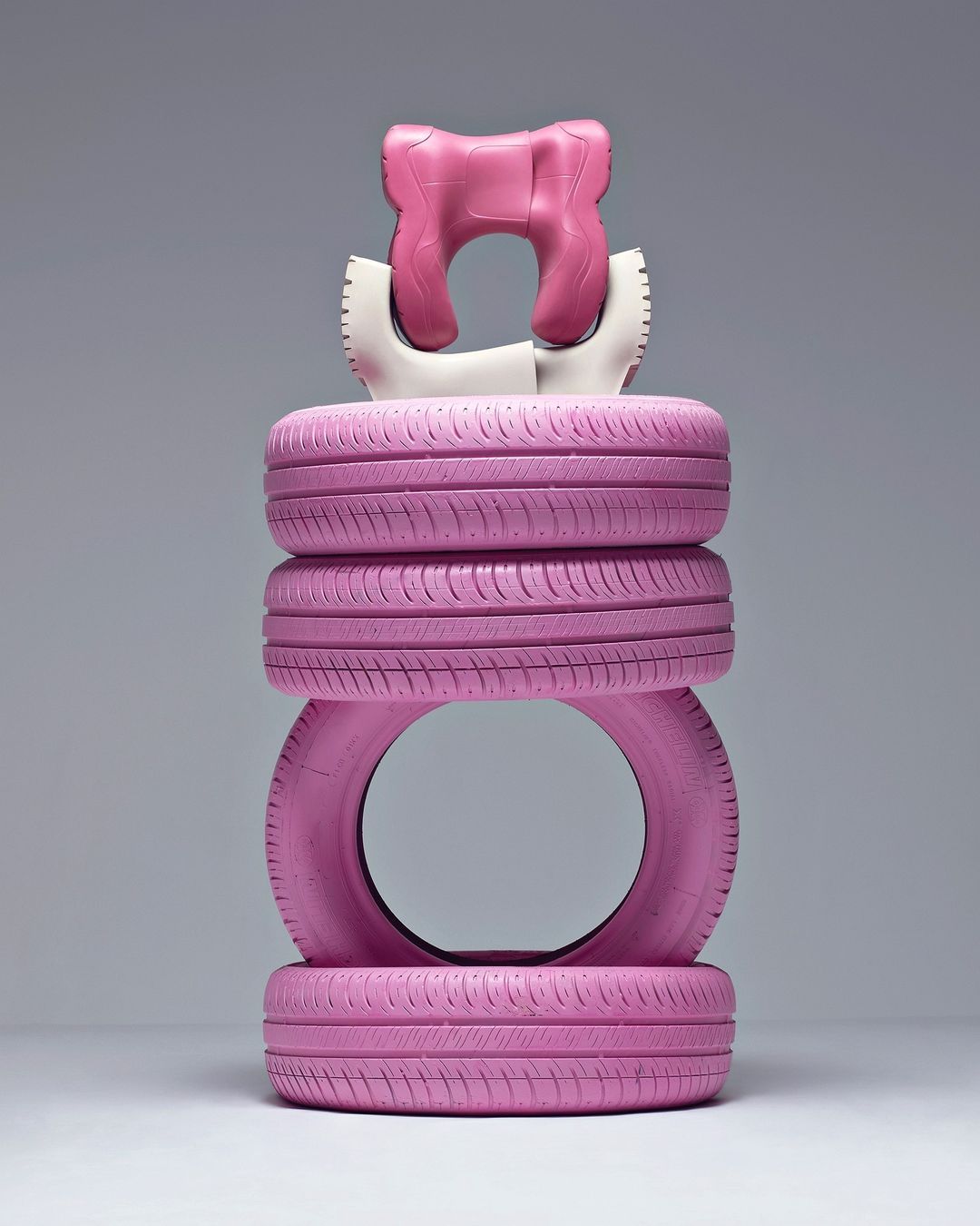 Tower of pink car tires and a pink-and-white neck pillow are vertically stacked against a grey background in a studio shot.