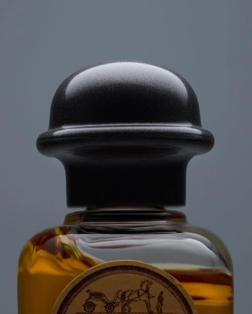 Close-up of a perfume bottle with a black cap and amber liquid against a grey background, showcasing its design and color.