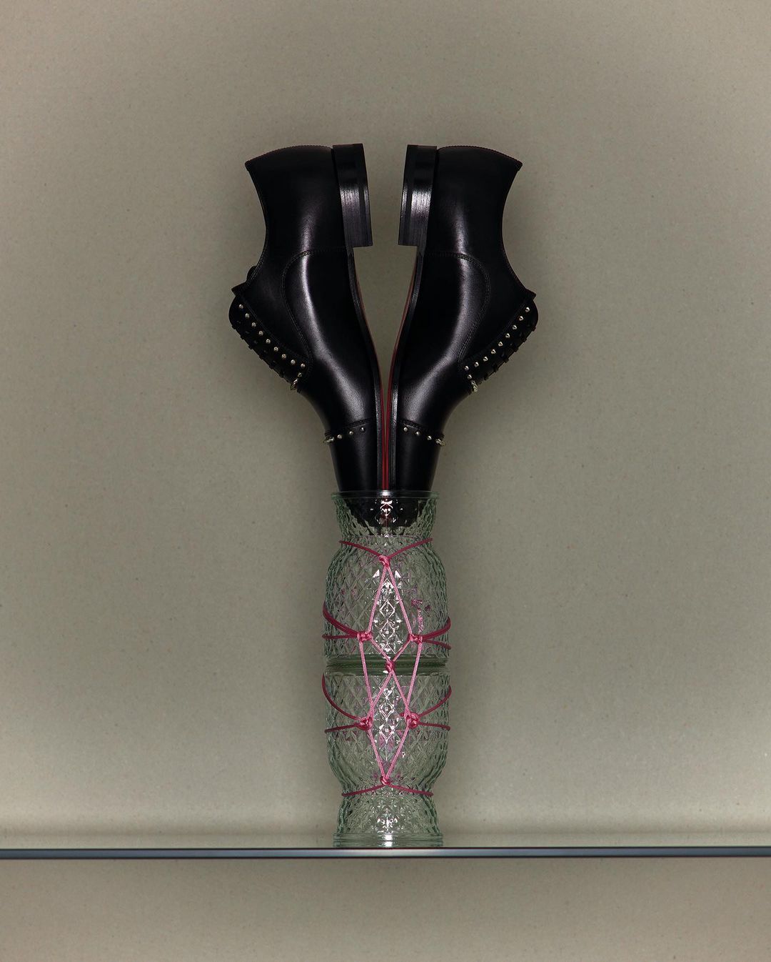 Pair of black leather dress shoes placed inside a clear glass vase wrapped with a pink string, displayed on a clear glass shelf.