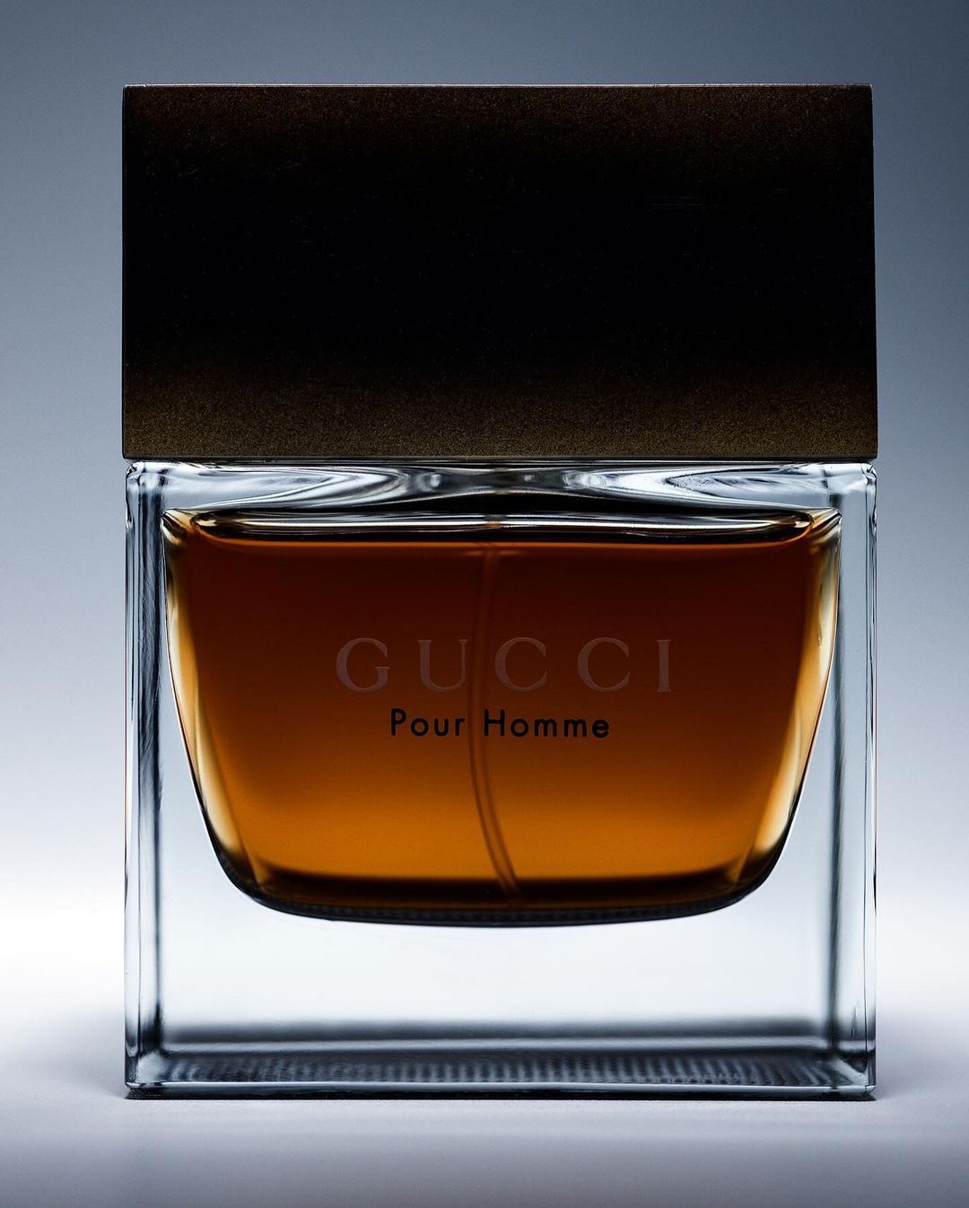 Elegant product shot of a glass bottle of Gucci Pour Homme cologne with a dark brown cap, centered on a pale gray background.