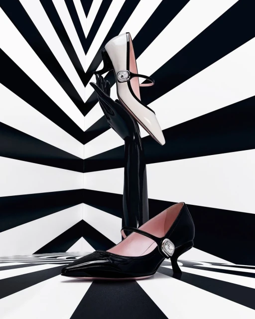 Black and white studio shot of two Mary Jane heels, one black and one white, posed on a black stand against striped background.