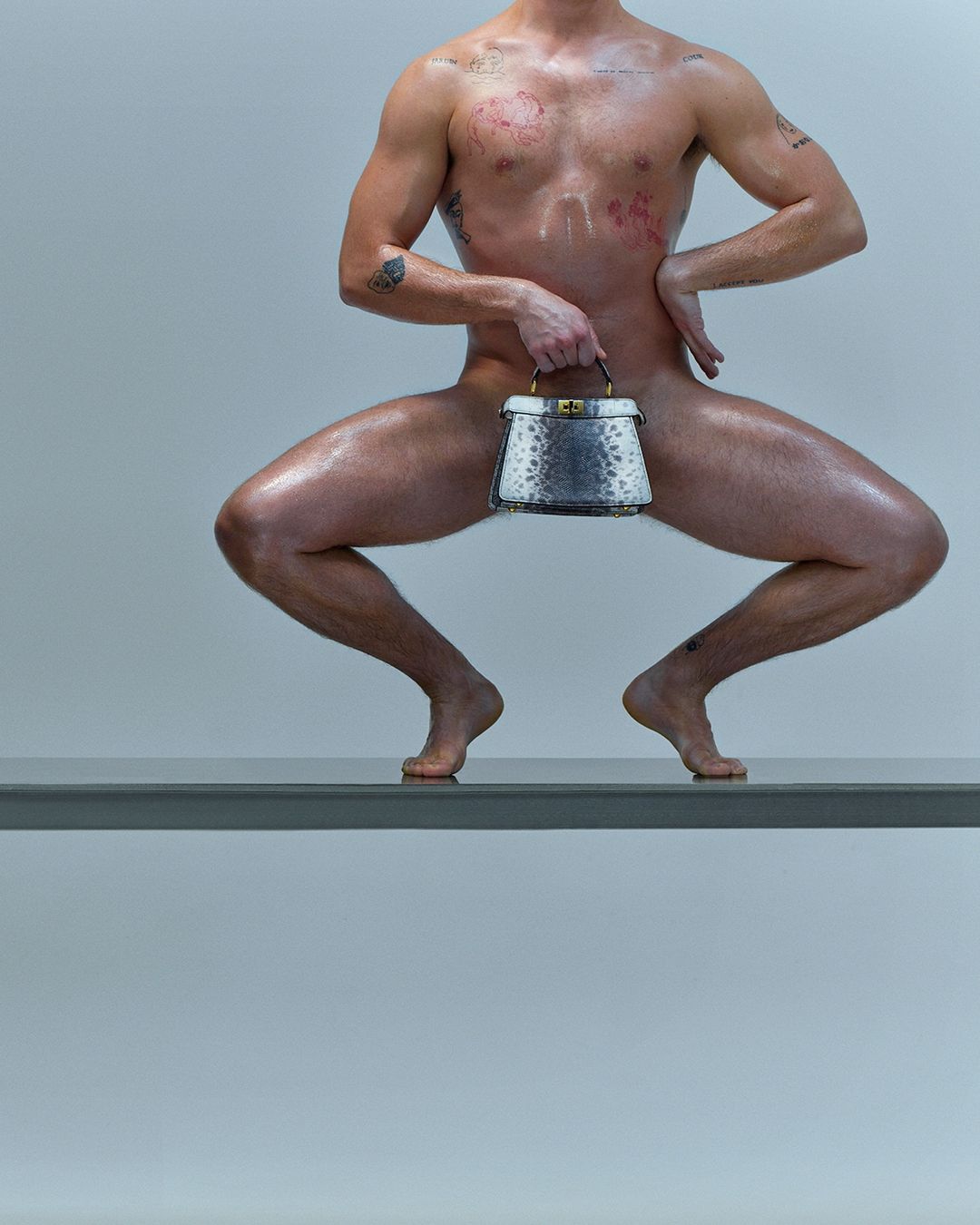 Shirtless man poses on a ledge with a handbag. He has tattoos and an athletic physique against a gradient gray background.