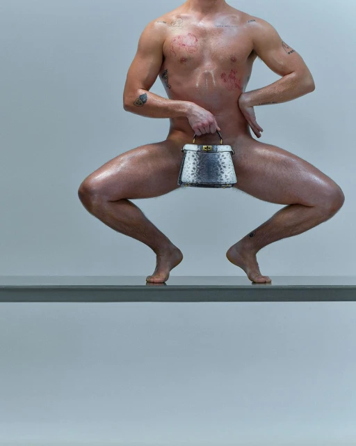 Shirtless man poses on a ledge with a handbag. He has tattoos and an athletic physique against a gradient gray background.